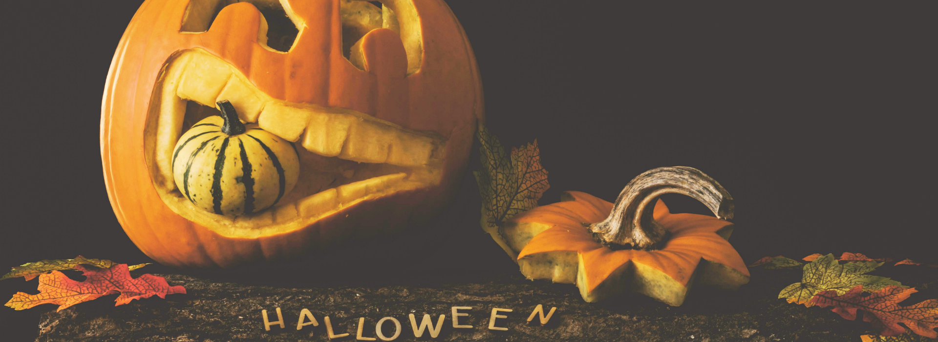Citrouille sculptée d’Halloween avec un sourire amusant tenant une petite courge dans sa bouche, accompagnée de feuilles d’automne et du mot « HALLOWEEN » formé sur le sol.